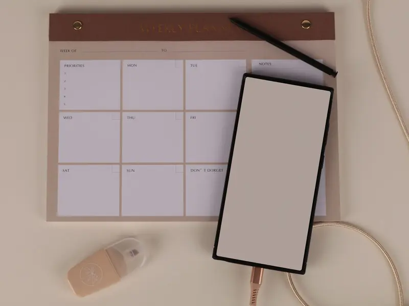 Planning hebdomadaire vierge sur un bureau beige, avec un smartphone et un stylo.